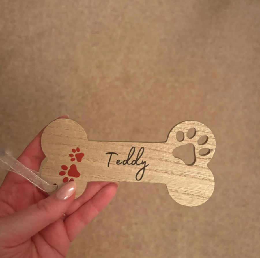 A hand holding a personalized wooden bone-shaped plaque engraved with the name 'Teddy' and decorated with red paw prints, against a beige carpeted background.